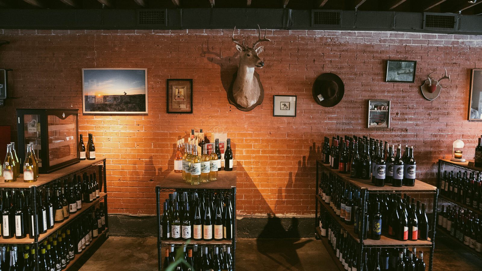 The wine room at Goodnight Cowboy, bottles on black racks against a red brick wall beneath a mounted deer