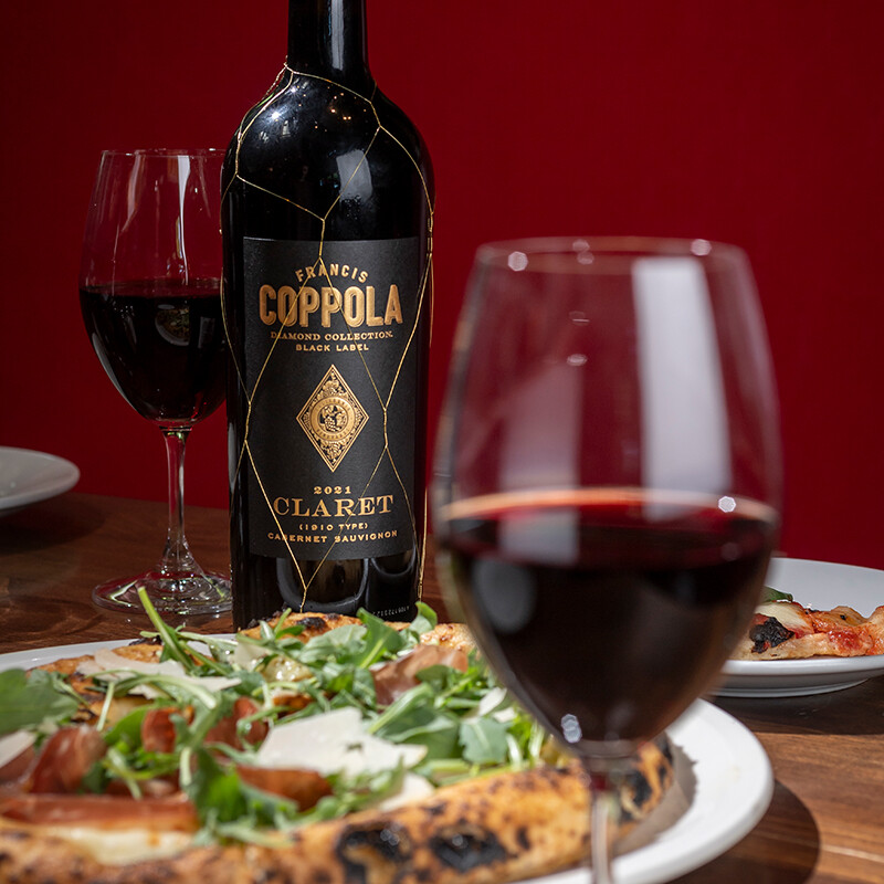 A bottle of Francis Ford Coppola Diamond Collection Black Label Claret in gold netting, standing on a wooden table behind a fresh pizza topped with arugula and prosciutto, with a glass of red wine in the foreground.