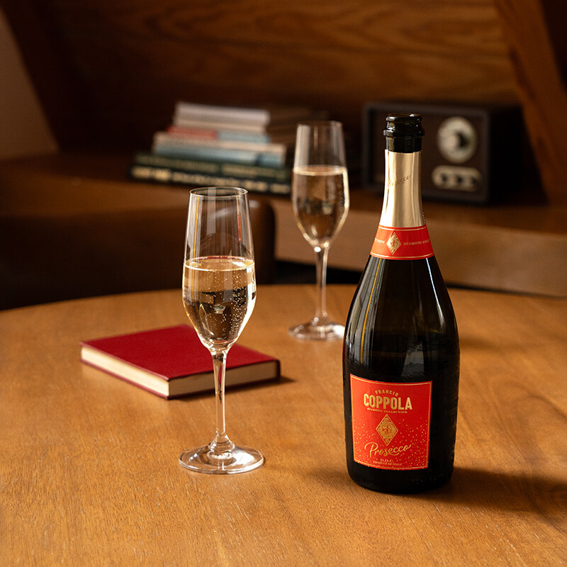 A bottle of Francis Ford Coppola Diamond Collection Prosecco with a red label standing on a wooden table next to a small red journal and two filled champagne flutes.