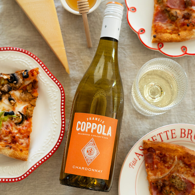 An overhead view of a Francis Ford Coppola Diamond Collection Chardonnay bottle with a bright orange label, arranged on a table runner alongside slices of pizza, a glass of white wine, a wedge of cheese, and a honey jar.
